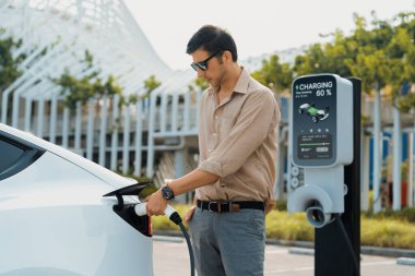 Genç adam, şehrin ticari otoparkındaki şarj istasyonundan elektrikli araba aküsünü şarj etmek için EV şarj aleti koymuş. Sürdürülebilir çevre dostu şehir yolculuğu için şarj edilebilir EV araba. Yararlı