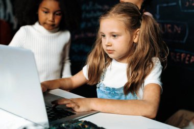 Beyaz önlüklü sarışın kız BTMM sınıfında kendine güvenen dizüstü bilgisayar kullanarak robot teknolojisini öğreniyor. Beyaz süveter giyen kız meraklı gözlerle kodlamasına bakıyor. Etkinlik.