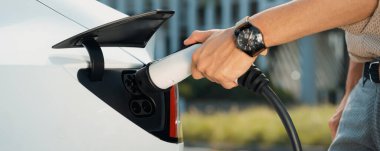 Genç adam şehrin ticari otoparkındaki şarj istasyonundan elektrikli araba aküsünü şarj ediyor. Sürdürülebilir çevre dostu şehir yolculuğu için şarj edilebilir EV araba. Panorama Çıkarı