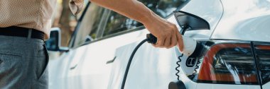 Genç adam yeşil şehir parkındaki şarj istasyonundan elektrikli araba aküsünü şarj ediyor. Sürdürülebilir çevre dostu şehir yolculuğu için şarj edilebilir EV araba. Panorama Çıkarı
