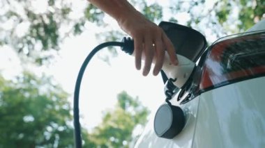 Yavaş çekim adamı, yaz mevsiminde açık yeşil yay yeşilliğindeki şarj istasyonundan elektrikli araba aküsünü şarj etmek için EV şarj aleti taktı. Yeniden şarj edilebilir elektrikli araç için sürdürülebilir enerji