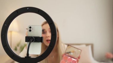Sosyal medya için güzellik ve kozmetik video içeriği hazırlayan genç bir kadın. Güzellik blogcusu kamera ve ışık halkası kullanarak seyirciye ve takipçiye nasıl göz farı uygulanacağını gösteriyor. Blithe