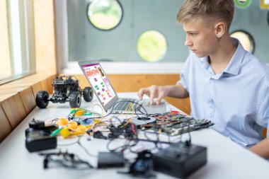 Liseli çocuk ekranda kod izliyor ve STEM dersinde teknoloji öğrenirken bilgisayarın dokunmatik pedini kullanıyor. Masaya dizüstü bilgisayar, kumanda, elektrik kablosu, şarj aleti ve robot araç koyun. Öğretim.