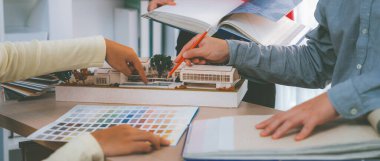 Proje yöneticisi toplantı masasında ev modeliyle ana tema rengini seçerken profesyonel iç tasarım ekibinin kırpılmış görüntüsü proje için renk ve malzeme kullanımı hakkında tartışır. Değiştirilmiş.