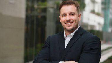 Kafkasyalı iş adamının kollarını kavuşturup şehir merkezindeki kameraya bakması. Proje yöneticisi yeşil şehirde takım elbise giyerken kendine güvenle ayakta duruyor. Yeşil çevre. Urbane.