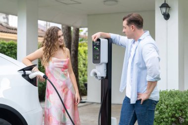 Çevre dostu, mutlu ve sevimli çift EV şarj istasyonundan elektrikli aracını şarj ediyor. EV araba teknolojisi gelecekteki sürdürülebilirlik için kullanılıyor. Eşzamanlama