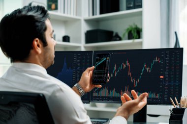 Akıllı telefondan akıllı telefon arayan tüccar işadamı, bilgisayardaki dinamik borsa yatırım ekranını analiz ediyor ve modern merkez ofisteki en kârlı finansal teknoloji pazarına yatırım yapıyor. Tahmin et..