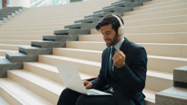 Kendine güveni tam bir iş adamı başarılı iş fikrini müzik dinlerken kutluyor. Happy yönetici projesi kulaklık ve takım elbise giyerken dizüstü bilgisayarla çalışıyor. Yatırımcı merdivenlerde oturuyor. Sevinçli..