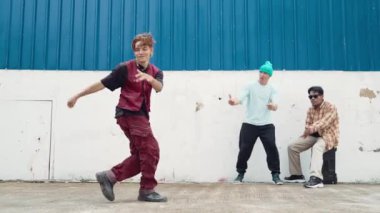 Genç Asyalı dansçı hip hop tarzında dans ediyor ve çok kültürlü arkadaşları duvarın arkasında tezahürat yapıyor. Çekici yakışıklı adam mavi arka planda kollarını uzatıyor. Açık hava sporu 2024 hiphop..