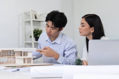 Profesyonel Asyalı erkek mimar ev modelinin uzunluğunu ölçmek için cetvel kullanırken, beyaz tenli genç meslektaşı ev modeliyle toplantı masasındaki verileri analiz etmek için dizüstü bilgisayar kullanıyor. Tertemiz..