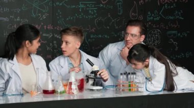 Çok kültürlü bir öğrenci mikroskop altında incelerken güzel bir kız da numuneyi inceliyor. BTMM sınıfında karatahta bilim teorisi deneyi yapan liseli çocuklara bakan öğretmen. Öğretim
