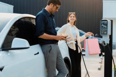 Alışveriş çantası tutan genç çift, alışveriş merkezinin otoparkındaki şarj istasyonundan EV araba aküsünü şarj etmek için akıllı telefon kullanıyor. Mutlu çift eco arabasıyla alışverişe gider. Yararlı