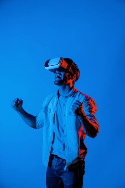 Profesyonel oyuncu, renkli neon ışıklı arka planda VR gözlük takarken oyunu kazanmasını kutlamak için elini havaya kaldırdı. Beyaz adam teknolojik yenilikleri kullanıyor ve giriyor. Sapma.