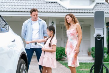 Mutlu küçük kız, ailesinin EV şarj istasyonundan elektrikli aracını şarj etmesine yardım ederken çevre dostu ve enerji sürdürülebilirliğini öğreniyor. EV araba ve modern aile kavramı. Eşzamanlama