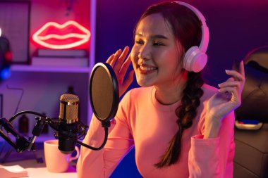 Genç ve güzel Asyalı yayıncının sunucu kanalı, dinleyicilerle sosyal medyada müzik dinliyor, pastel kulaklık takıyor, kapalı alanda neon ışıklı stüdyoda şarkı söylemek için mikrofon kullanıyor. Taktik.