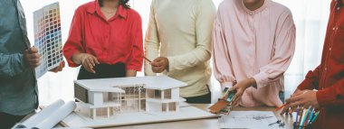 Profesyonel mimar takımının ev rengi seçimi hakkında beyin fırtınası yaparken, mimar da plan ve ev modelinin olduğu masadaki taslak belgeye yazı yazıyor. Değiştirilmiş.