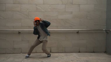 Asyalı akıllı hipster, kulaklık ve şık kıyafetler giyip hip hop şarkıları dinlerken gri arka planlı enerjik break dans gösterisi yapıyor. Açık hava sporu 2024. Hip hop hareketi. Yumuşak bir şekilde..