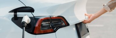 Otoparktaki şarj istasyonunun pilini şarj ederken genç bir kadın bagajı açıyor. Sürdürülebilir çevre dostu seyahat için alternatif enerji ve şarj edilebilir elektrikli araç.