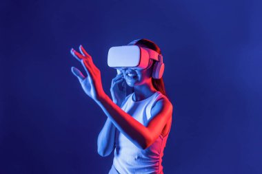 Çevresi siberpunk neon ışıklarıyla çevrili zeki bir kadın metaevrene bağlı VR kulaklık takıyor, gelecekçi siber uzay topluluğu teknolojisi. Sanal objeye el ile dokunan kadın. Halüsinasyon.