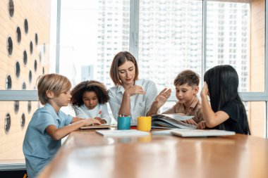 Öğretmen sınıftaki kitaplardan ders çıkarmayı öğretiyor, çocuklar mutlu. Bazıları öğretmenine soruyor, mavi gömlekli bir çocuk tek başına kitap okuyor ve bir kız resim çiziyor. Etkinlik.