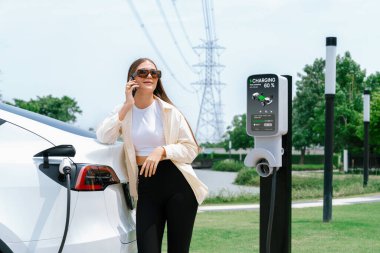EV araba aküsünü şarj eden genç bir kadın elektrik şebekesine bağlı elektrik santralinde çevre dostu araç kullanımı için elektrik endüstrisi olarak telefonla konuşurken. Yararlı