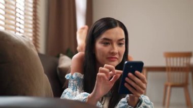 Kanepede yatan genç bir kadın internet bankacılığı için kredi kartıyla dizüstü bilgisayar kullanıyor. Evdeki online ödeme kapısından alışveriş yapıyor. Modern ve uygun online satın alma. Blithe