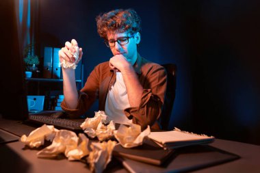 Aşırı çalışan projeyle uğraşan stresli yaratıcı yönetici buruşmuş kağıtların etrafını sararken gece vakti modern ofisteki gündelik gömlekle dizüstü bilgisayar ekranına e-posta göndermeyi bekliyor. Toplayıcı.