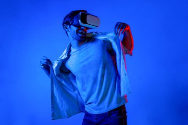 Yakışıklı, dans eden ve müziğe hareket eden bir adam. VR gözlüğü ve neon ışıklı arka plan kullanarak. Beyaz erkek oyuncu, teknolojik yenilikler giyerek pop müzik dinlerken ayakta duruyor. Sapma.