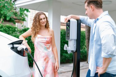 Çevre dostu, mutlu ve sevimli çift EV şarj istasyonundan elektrikli aracını şarj ediyor. EV araba teknolojisi gelecekteki sürdürülebilirlik için kullanılıyor. Eşzamanlama