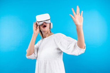 Mutlu kadın VR gözlüklerine bakıp mavi arka planda dururken şaşırdı ya da heyecanlandı. Beyaz pijamalı beyaz bir kız metaevrene bağlanmak için görsel gerçeklik gözlüğü kullanıyor. Kısıtlanma.