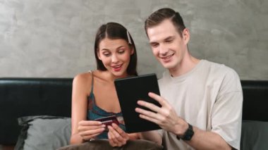 Genç çift, kredi kartıyla ödeme yapabilmek için internet üzerinden ödeme uygulaması ve dijital cüzdan kullanarak yatak odasında oturuyor. Mobil internet üzerinden alışveriş ve modern alımlar. Düzenle