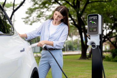 Açık yeşil şehir parkı manzaralı EV şarj istasyonundan genç kadın EV elektrikli araçları şarj ediyor. Çevre dostu şehir ulaşımı ve çevre dostu EV araba seyahati. Yükselt