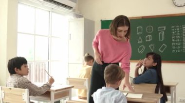 Zeki Asyalı öğrenci, ilkokulda öğretmen yürürken ve ödevlerini kontrol ederken matematik sorusunu sorarak elini kaldırdı. Öğretmen not defterini alırken soruyu cevaplıyor. Pedagoji.