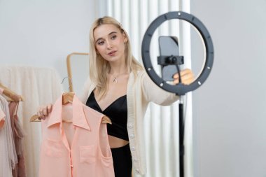 Genç sosyal medyum kadın moda videosu yapıyor. Blogcu kameraya ve ışık çemberine gülümserken, aynı zamanda ikna edici çevrimiçi kıyafetleri izleyicilere veya takipçilere vlog sattırıyor. Blithe