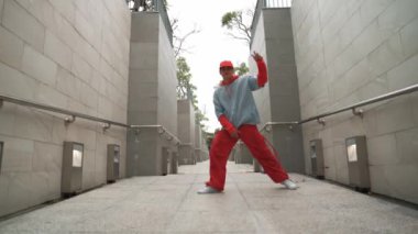 Profesyonel hipster koridorda yürürken ayak sesleri çıkarıyor. Yetenekli sokak dansçısı B-boy dansını gösterirken kameraya komik bir hareket yapıyor. Modern yaşam tarzı. Açık hava sporu 2024. Yumuşak bir şekilde..