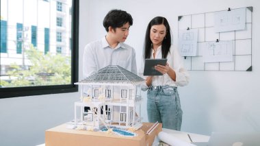 Güzel, genç, beyaz bir proje yöneticisi Asyalı mühendise, mühendislik masasında ev modelini ölçerken tablet kullanarak bina detaylarını anlattı. Tertemiz.