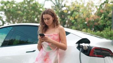 Genç bir kadın doğadaki EV şarj istasyonunda elektrik için akıllı telefon kullanıyor. Modern çevre ve EV araçlı sürdürülebilir otomobil taşıma yaşam tarzı. Titiz