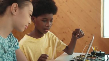 Afrika 'lı bir erkek ve beyaz bir kızın günlük giysiler içinde BTMM teknoloji sınıfında dizüstü bilgisayar ve elektronik ekipmanlarla kod yazma ya da programlama sisteminde birlikte çalıştıklarını gösteren site görüntüsü. Öğretim