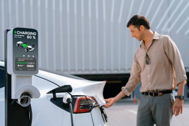 Genç adam, şehrin ticari otoparkındaki şarj istasyonundan elektrikli araba aküsünü şarj etmek için EV şarj aleti koymuş. Sürdürülebilir çevre dostu şehir yolculuğu için şarj edilebilir EV araba. Yararlı