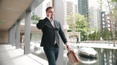 Modern koridorda yürürken telefonla konuşan mutlu proje yöneticisi. Parkta ya da yeşil şehirde yürürken finansal stratejiyi planlamak için pazarlama ekibini arayarak telefonu kullanıyor. Urbane.