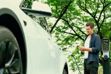 Ulusal Park veya Yaz Ormanı 'ndaki tatil gezisi sırasında EV şarj istasyonundan elektrik aküsü almak için akıllı telefon internet bankacılık uygulaması kullanan bir adam. Yükselt