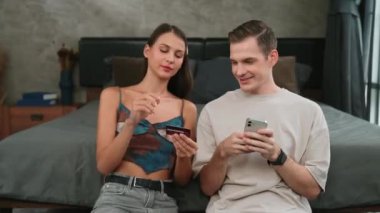 Genç çift, kredi kartıyla ödemek için internet üzerinden ödeme uygulaması ve dijital cüzdan kullanarak yatak odasında oturuyor. Mobil internet üzerinden alışveriş ve modern alımlar. Düzenle