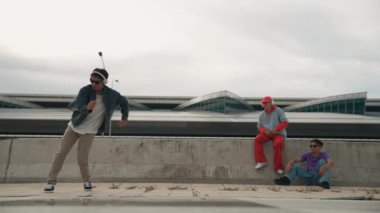 Yakışıklı break dansçı, Asyalı arkadaşının tezahürat yaptığı serbest stil hareketinden hoşlanır. Hippi kulaklıktan müzik dinler ve kameraya bakar. Sokak dansı. Açık hava sporu 2024. Yumuşak bir şekilde..