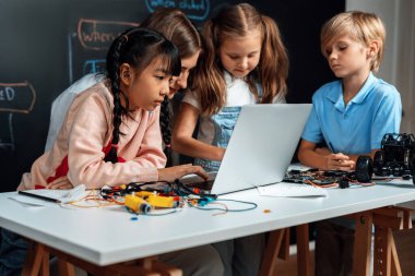 Uzaktan kumandalı araba programlamayı öğrenen yetenekli çocuklar. Pembe elbiseli liseli kız kodlama için dizüstü bilgisayar kullanıyor. Öğretmen onu izlerken diğer çocuk kodunu izliyor. Anakart ve elektrik kablosu da masada. Etkinlik.