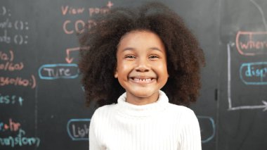 Genç Afrikalı bir kızın karatahtaya bakarken kameraya bakışı. BTMM teknoloji sınıfında arkada yazılı kamera sistemi koduna gülümseyen bir öğrenci. Etkinlik.