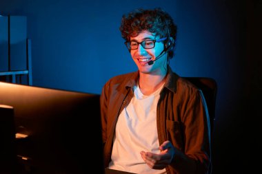 Genç yaratıcı iş adamı, bilgisayar ekranında dinleyicilerle iletişim kuruyor ya da takım üyesiyle görüşüyor. Neon Light Modern Ev Ofisi 'nde kulaklık takıyor. Gece vakti çalışma kavramı. Toplayıcı.