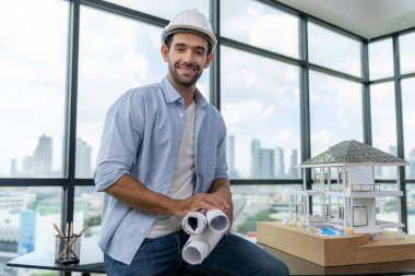 Modern ev projesini düşünen profesyonel mimar mühendisinin portresi güvenlik kaskı ve günlük kıyafetler giyerken. Akıllı iç mimar ev modelinin yanında proje planları tutuyor. İzleme.