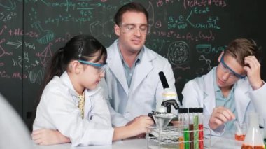 Zeki öğretmen, çeşitli öğrencilerin yaratıcı eğitim üzerine yazılmış bilim teorisiyle karatahta önünde deney yaparken test tüpü masanın üzerine yerleştirilirken mikroskop altında bakar.