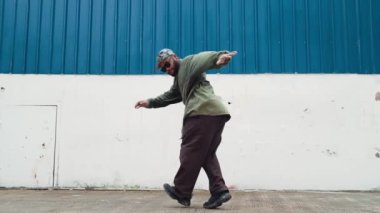 Sokak ortasında mavi duvarlı hippi dansçı ayak basamağı. Asyalı break dansçı kollarını açarken sokak dansı yapıyor. Hip hop koreografı enerjik bir dans sergiliyor. Açık hava sporu 2024 hiphop..