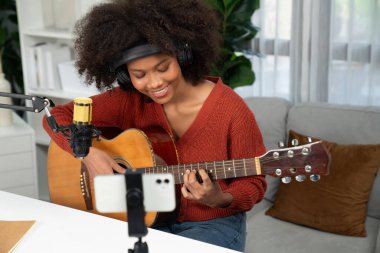 Genç Afrikalı Amerikalı müzisyenin sunucu kanalı gitar çalıyor, şarkı söylüyor, stüdyoda akıllı telefondan yayın yapıyor. Ekipman kulaklıkları dekorasyonu ve mikrofon kaydı. Tatlandırıcı.